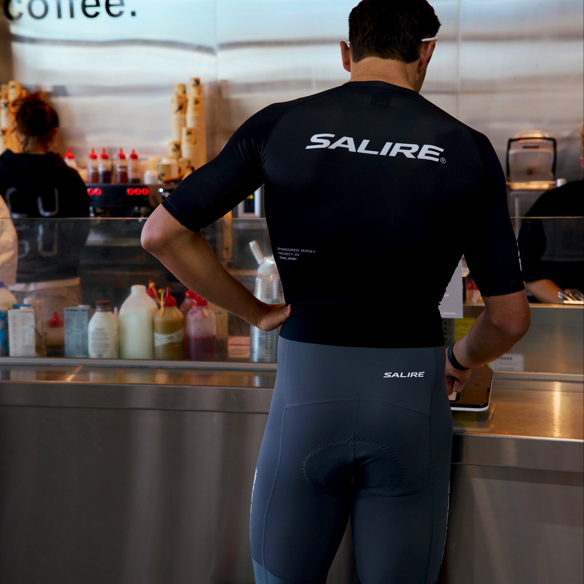 A man wearing a black Salire cycling jersey and grey bibs stands at a cafe