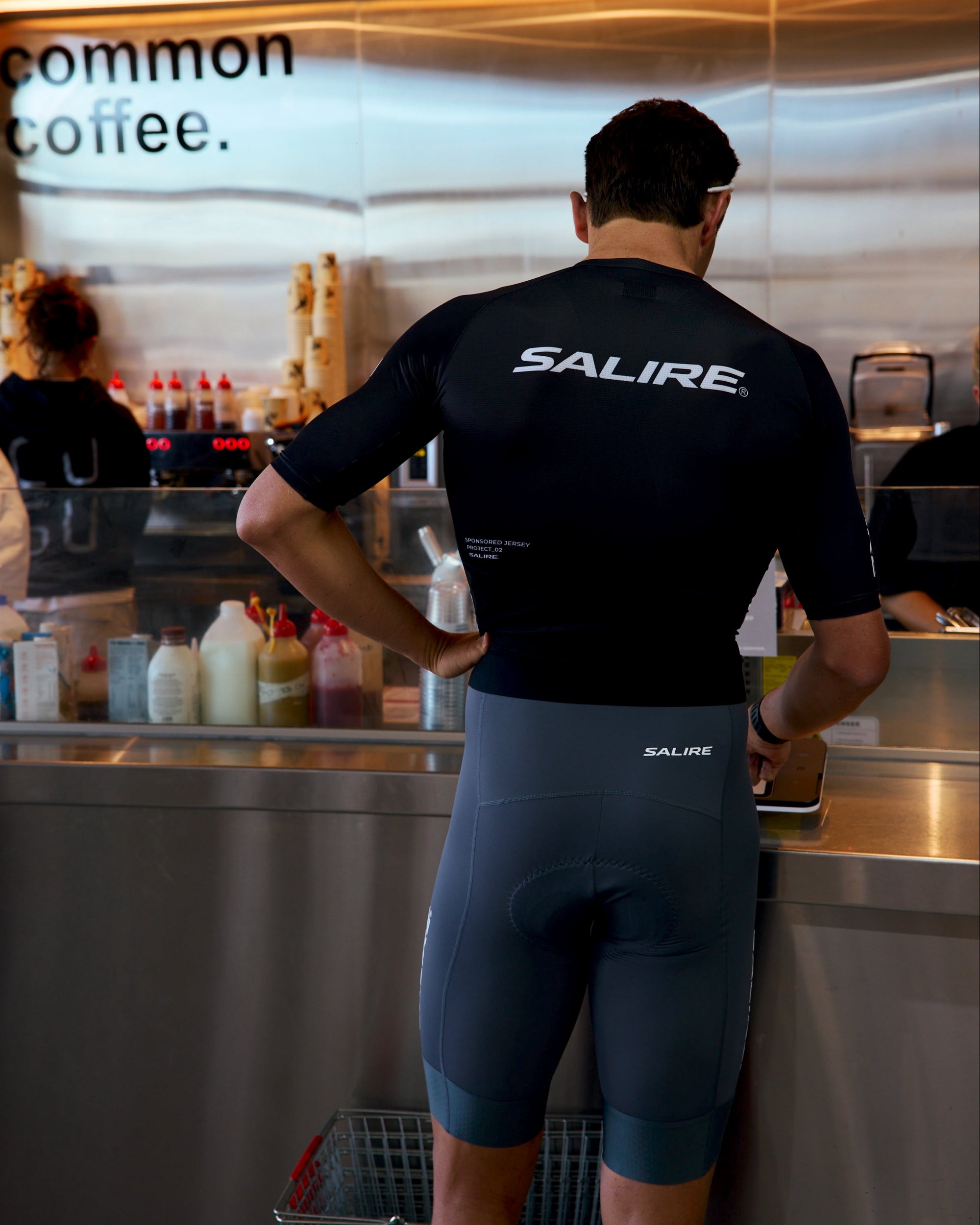 A man wearing a black Salire cycling jersey and grey bibs stands at a cafe